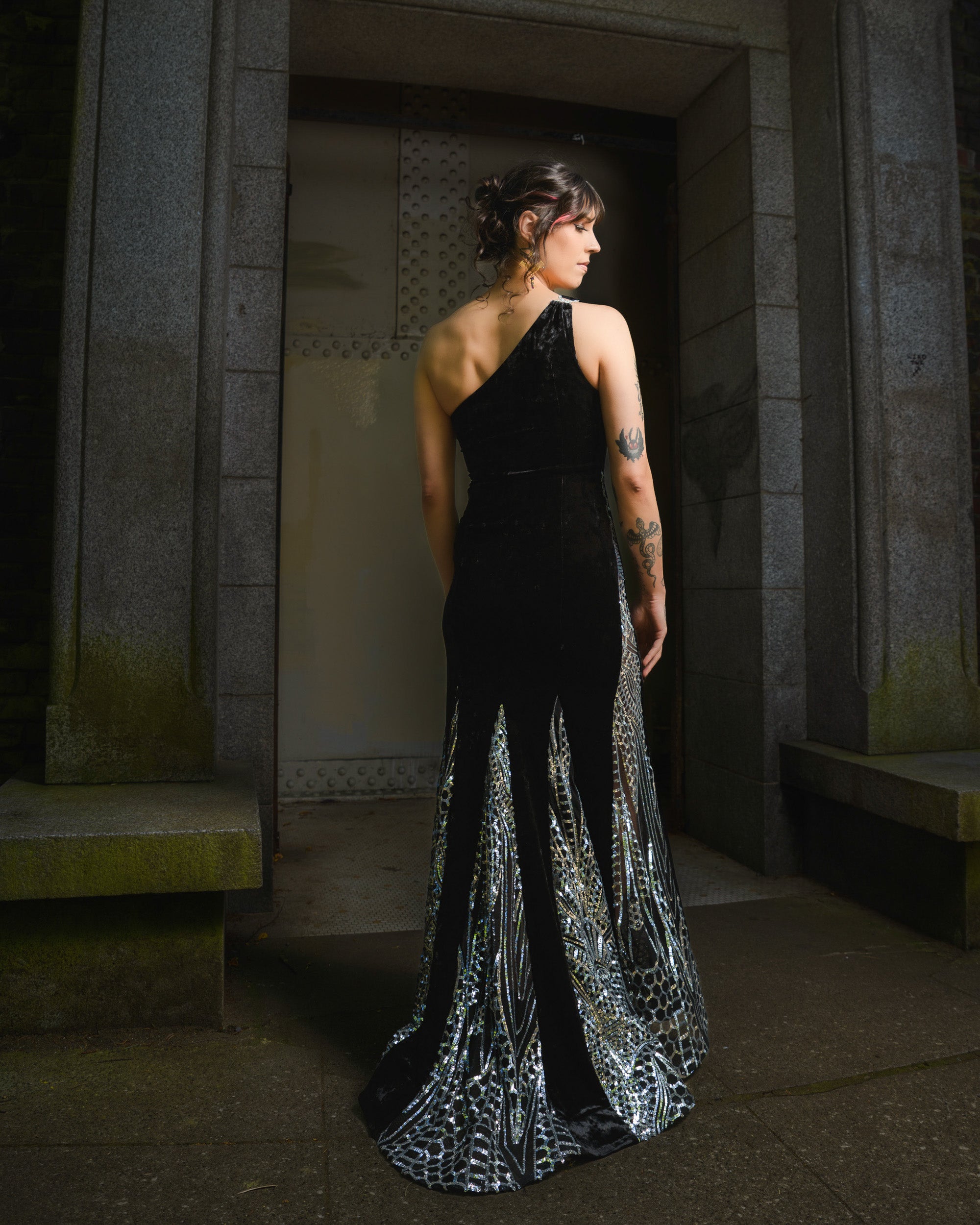 Woman in a black one-shoulder gown with silver detailing standing in a dimly lit hallway.  [model_size:M] [dress_size:L]  [editorial] [color_id:black]