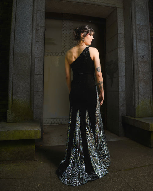 Woman in a black one-shoulder gown with silver detailing standing in a dimly lit hallway.  [model_size:M] [dress_size:L]  [editorial] [color_id:black]