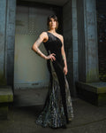 Woman in a black evening gown with intricate detailing standing in an urban setting. [model_size:M] [dress_size:L] [editorial] [color_id:black] [hero] 