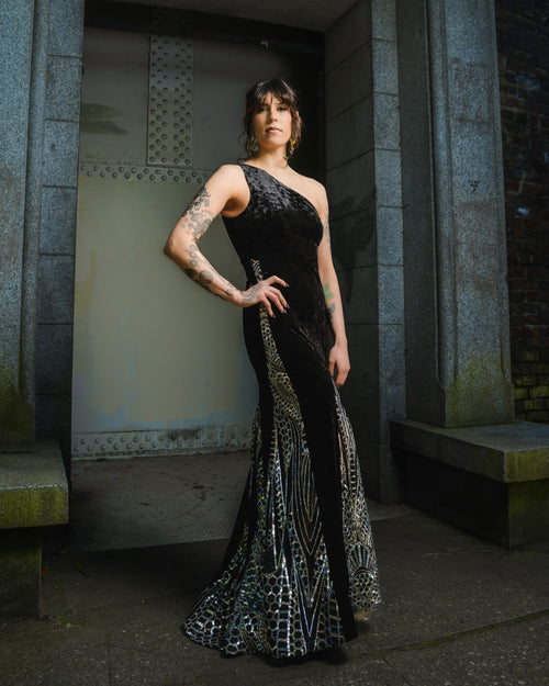 Woman in a black evening gown with intricate detailing standing in an urban setting. [model_size:M] [dress_size:L] [editorial] [color_id:black] [hero] 