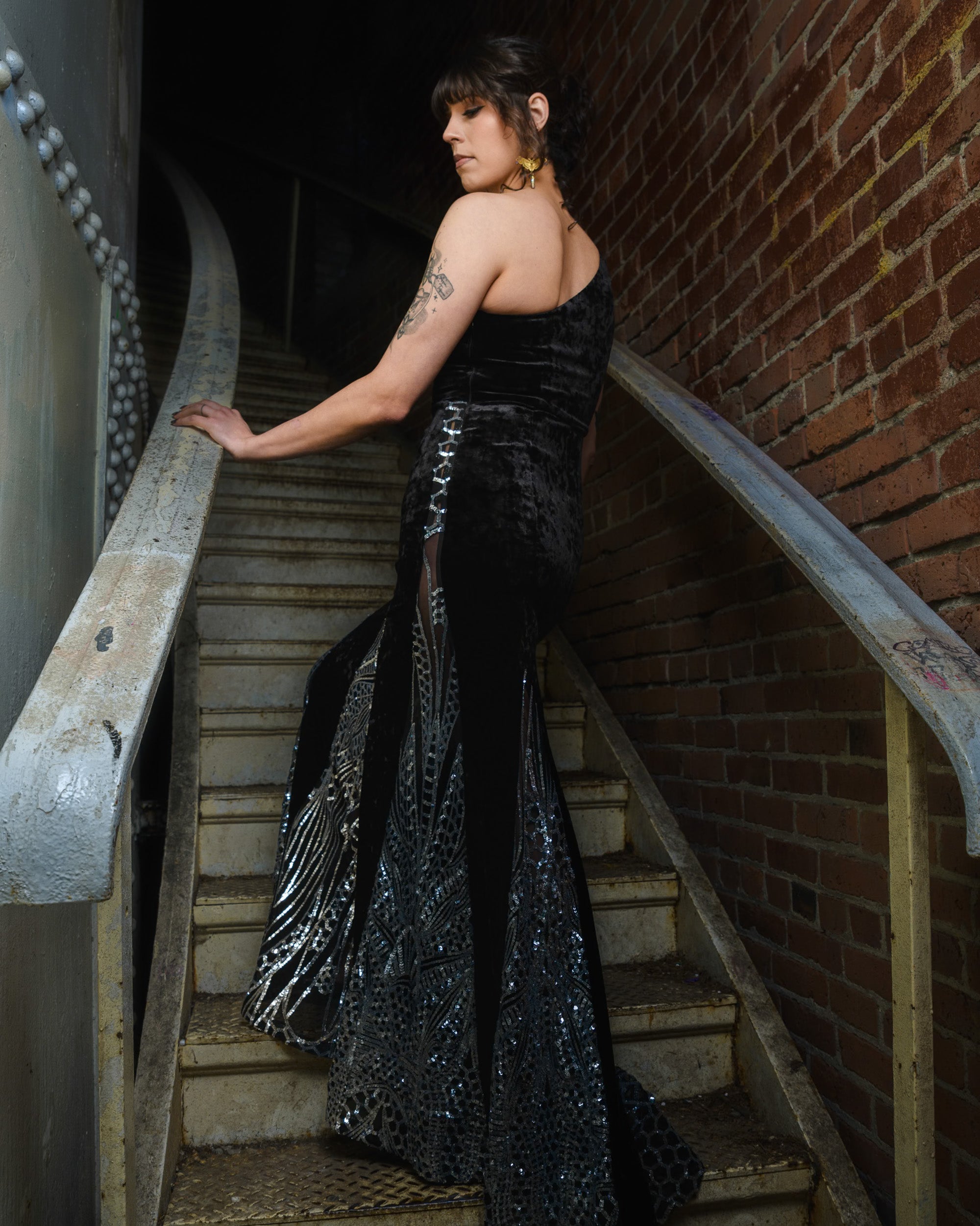 Woman in a black evening gown with silver accents standing on a staircase.  [model_size:M] [dress_size:L]  [editorial] [color_id:black]