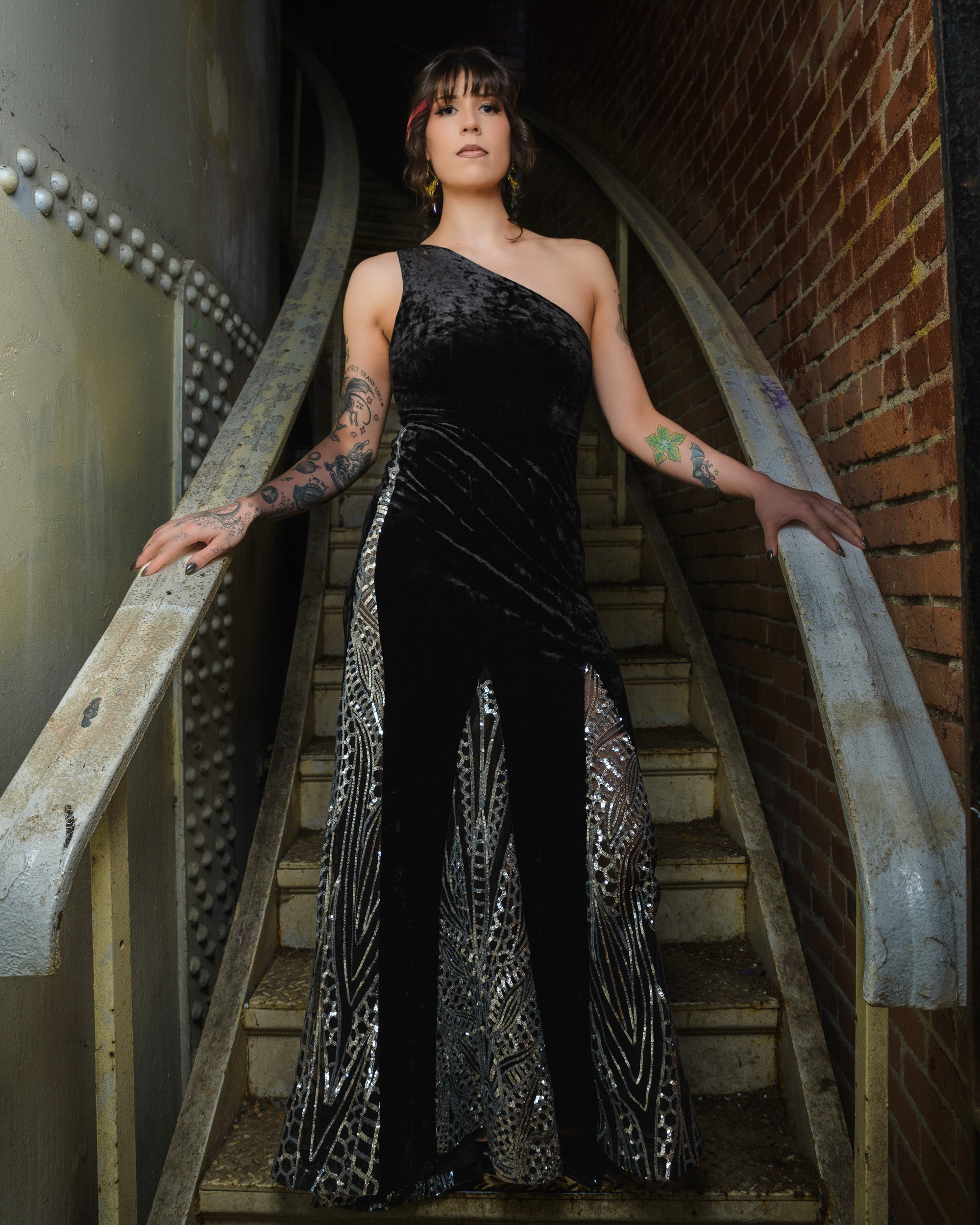 Woman in a black evening gown with silver embellishments standing on a staircase. [model_size:M]  [dress_size:L] [color_id:black] [editorial]