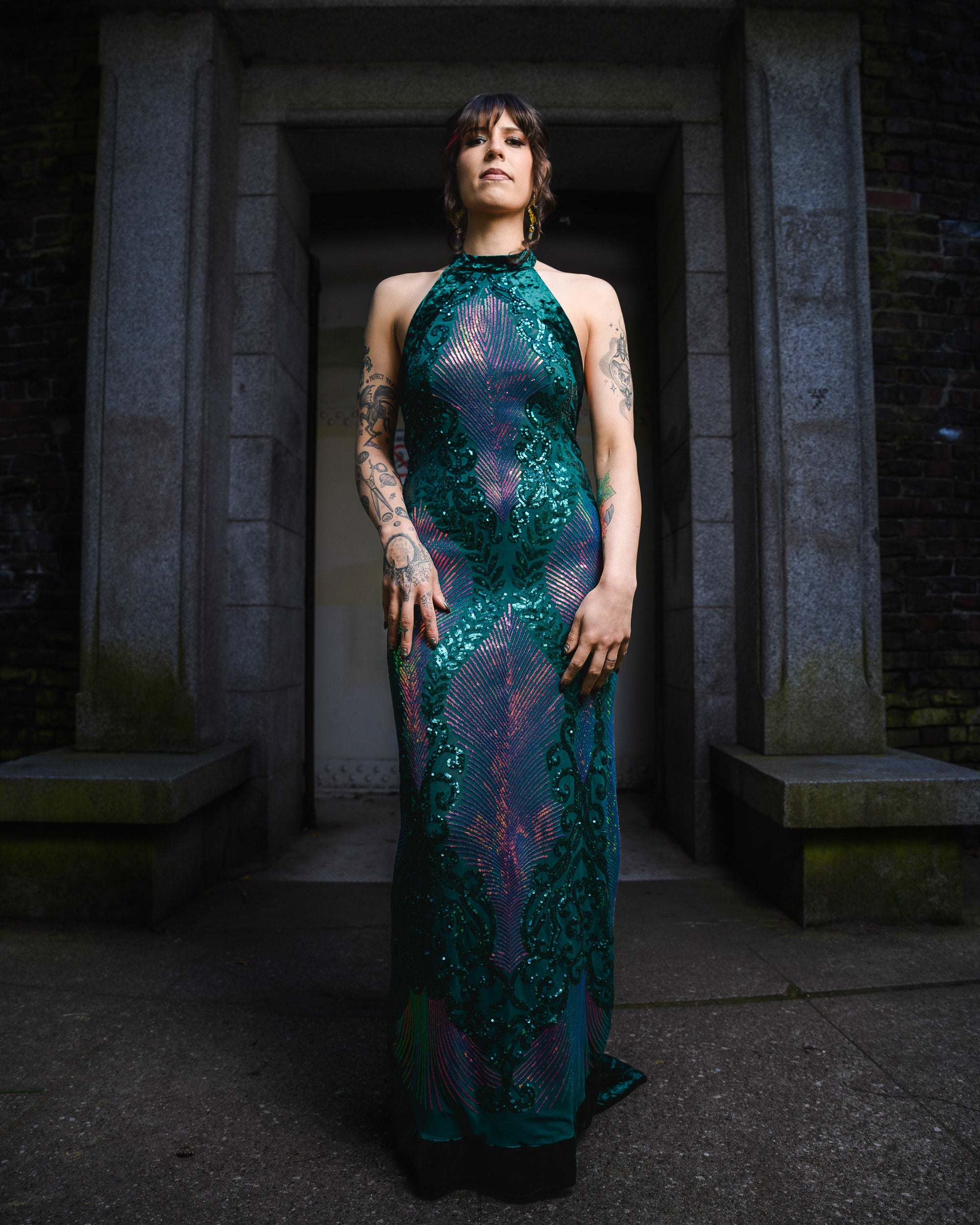 Woman in a green sequined dress standing in a dark, stone-walled corridor. [color_id:green-purple] [editorial] [hero]