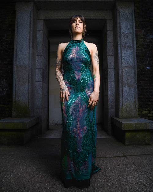 Woman in a green sequined dress standing in a dark, stone-walled corridor. [color_id:green-purple] [editorial] [hero]