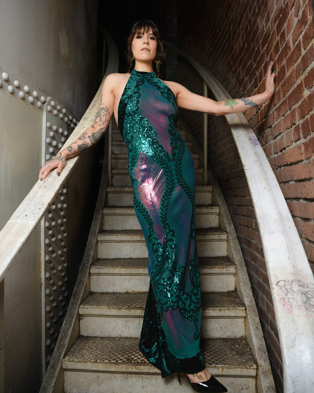 Woman in a green sequin dress standing on a staircase with a brick wall background [model_size:M] [dress_size:M] [color_id:green-purple] [editorial]