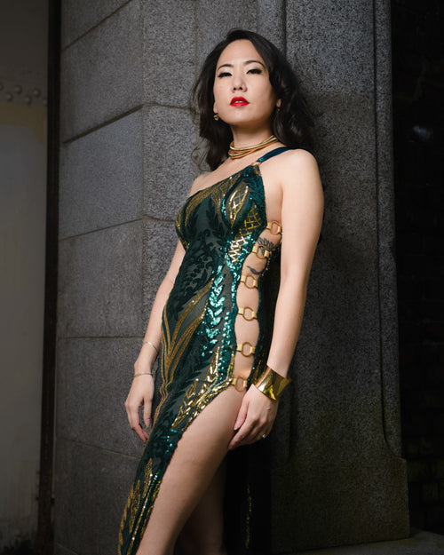 Woman in a green and gold dress standing against a gray wall.  [color_id:green-gold] [editorial]