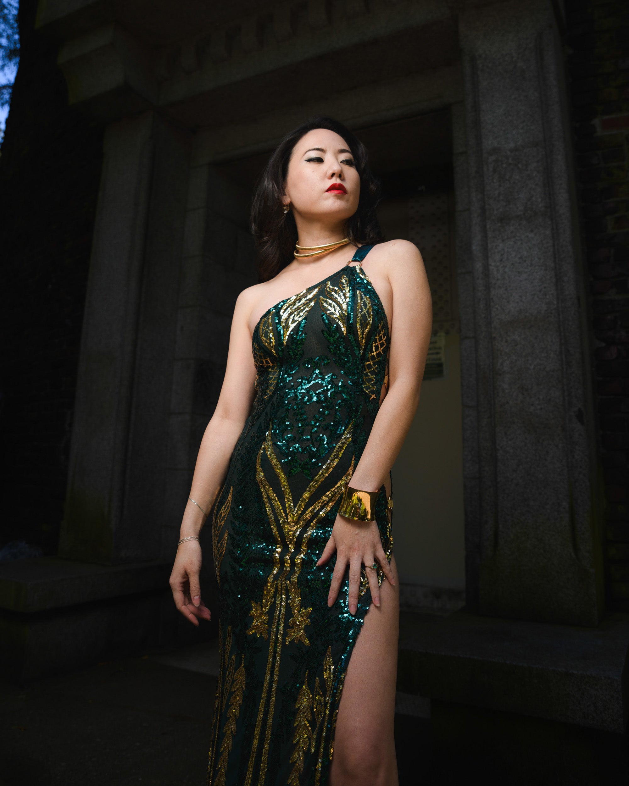 Woman in a green and gold sequin dress standing in a dark, architectural setting. [color_id:green] [editorial]