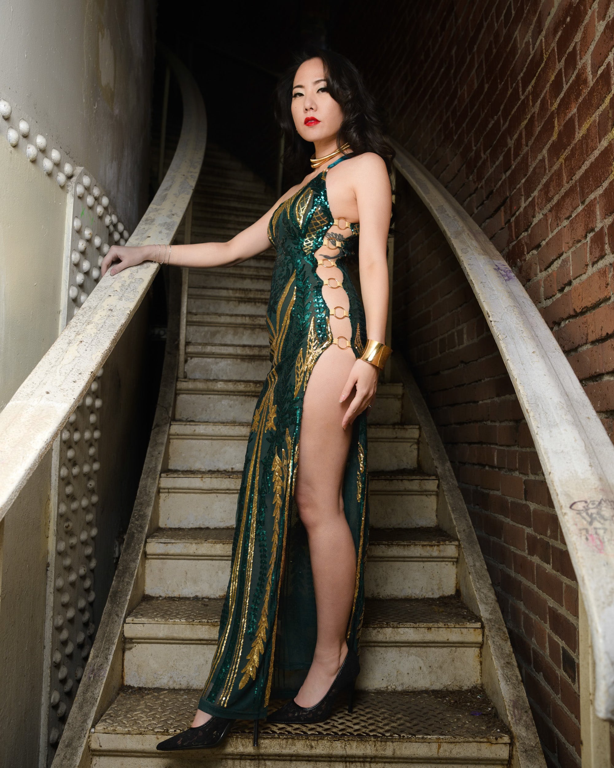 Woman in a green and gold dress standing on a staircase with a brick wall background   [color_id:green] [editorial] [hero] [bogus tag]