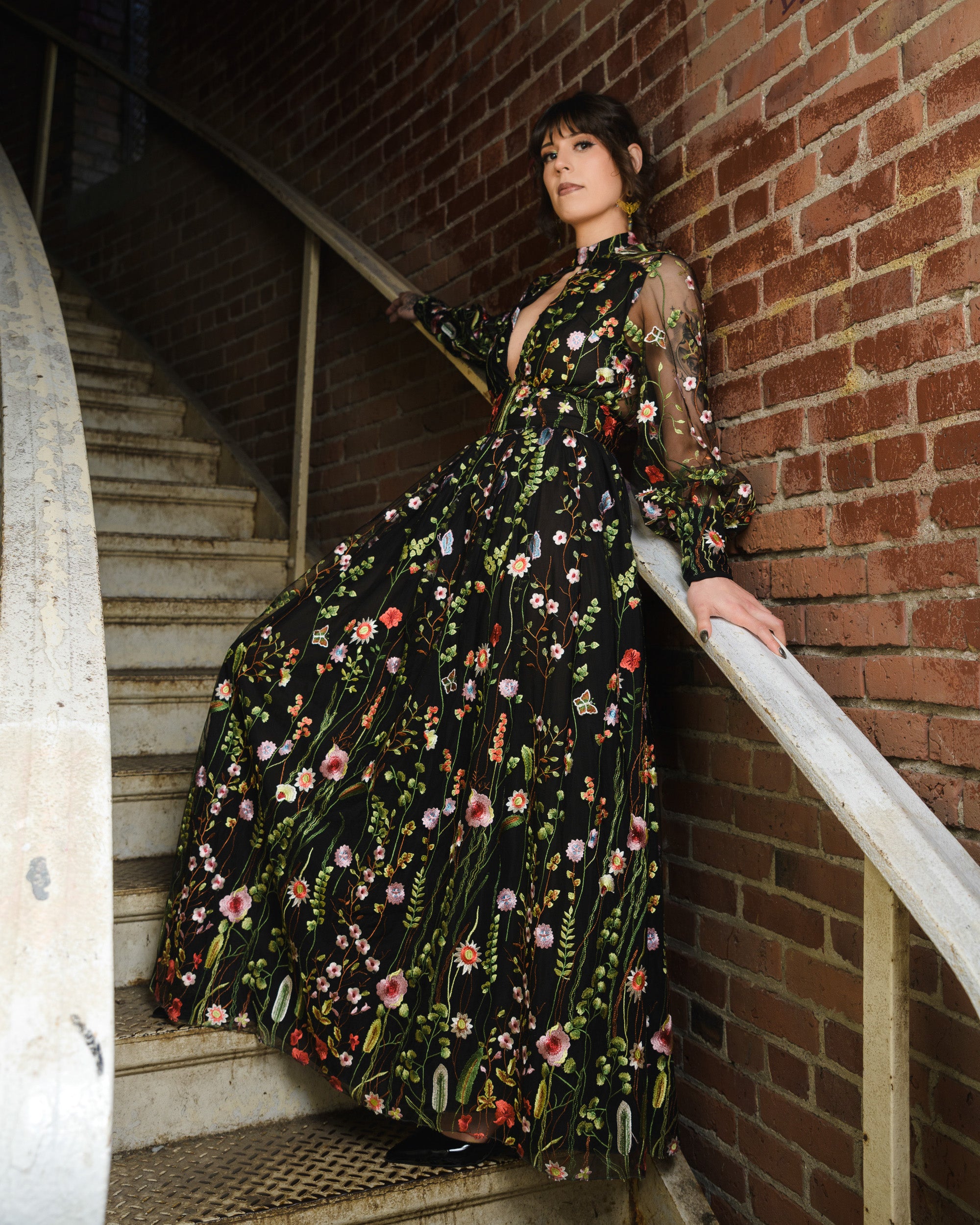 Woman in a floral dress standing on a staircase against a brick wall [color_id:black] [editorial] 