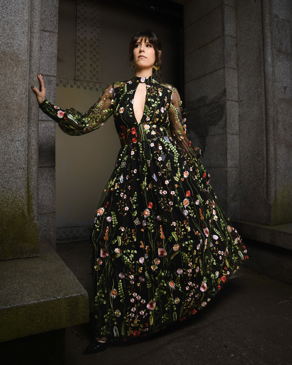 Woman in a floral dress standing in a dimly lit room with stone walls. [color_id:black] [editorial] [hero] [model_size:M] [dress_size:L]  
