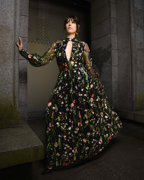 Woman in a floral dress standing in a dimly lit room with stone walls. [color_id:black] [editorial] [hero]