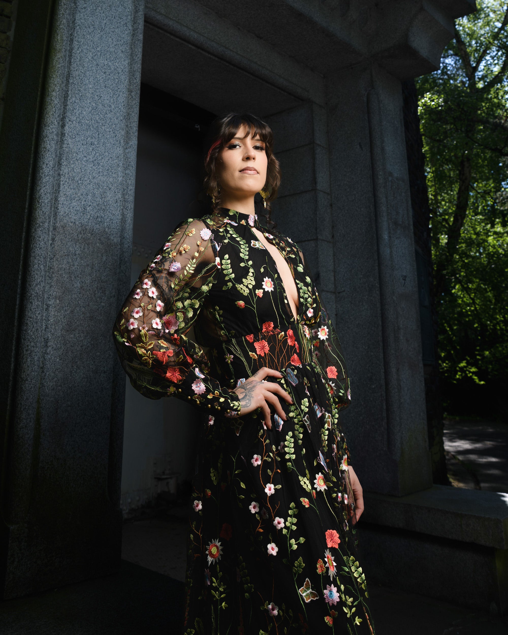 Woman in a floral dress standing in a dimly lit outdoor setting with trees in the background.  [color_id:black] [editorial] 
