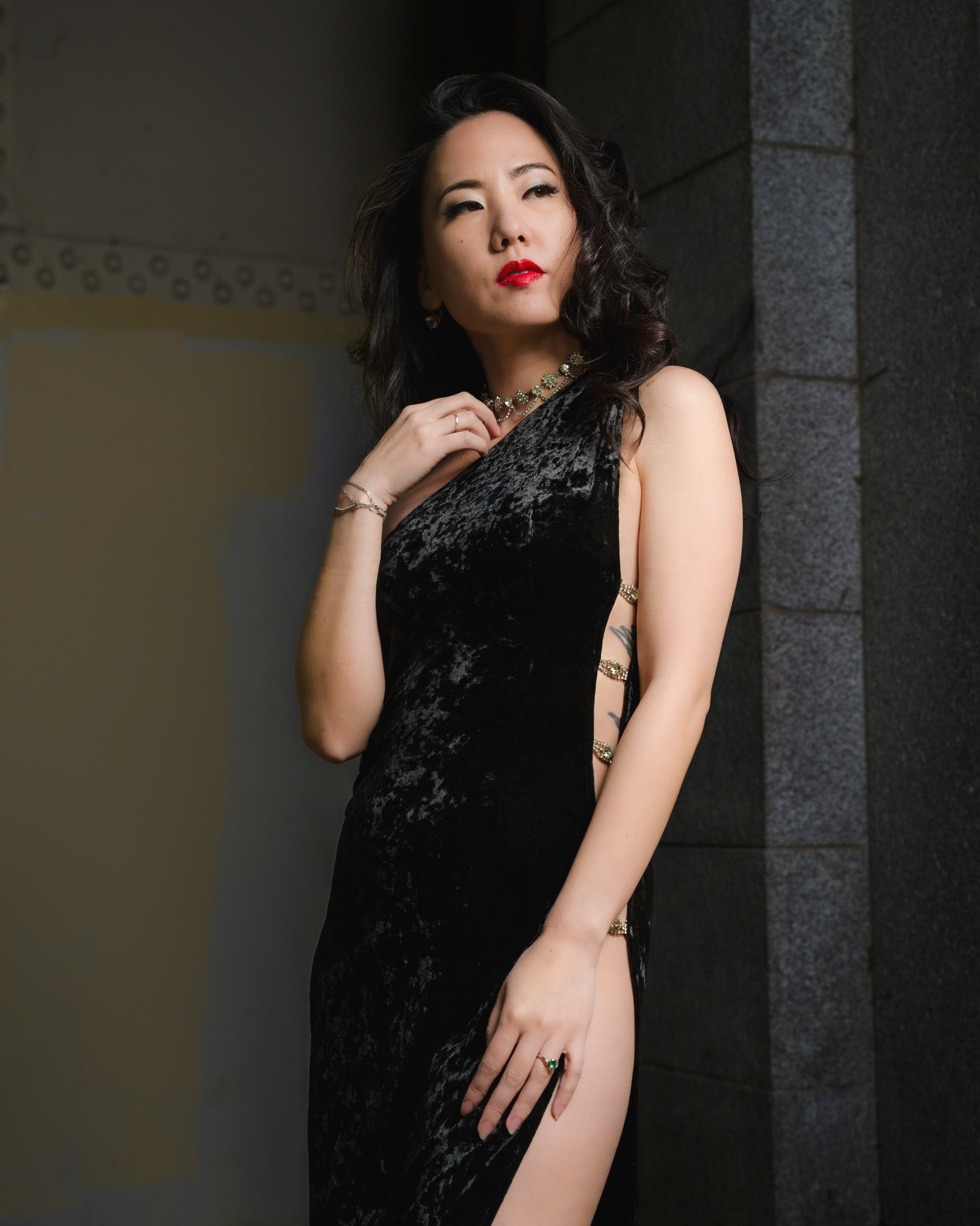Woman in a black dress standing against a dark wall. [color_id:gunmetal] [editorial] [hero]