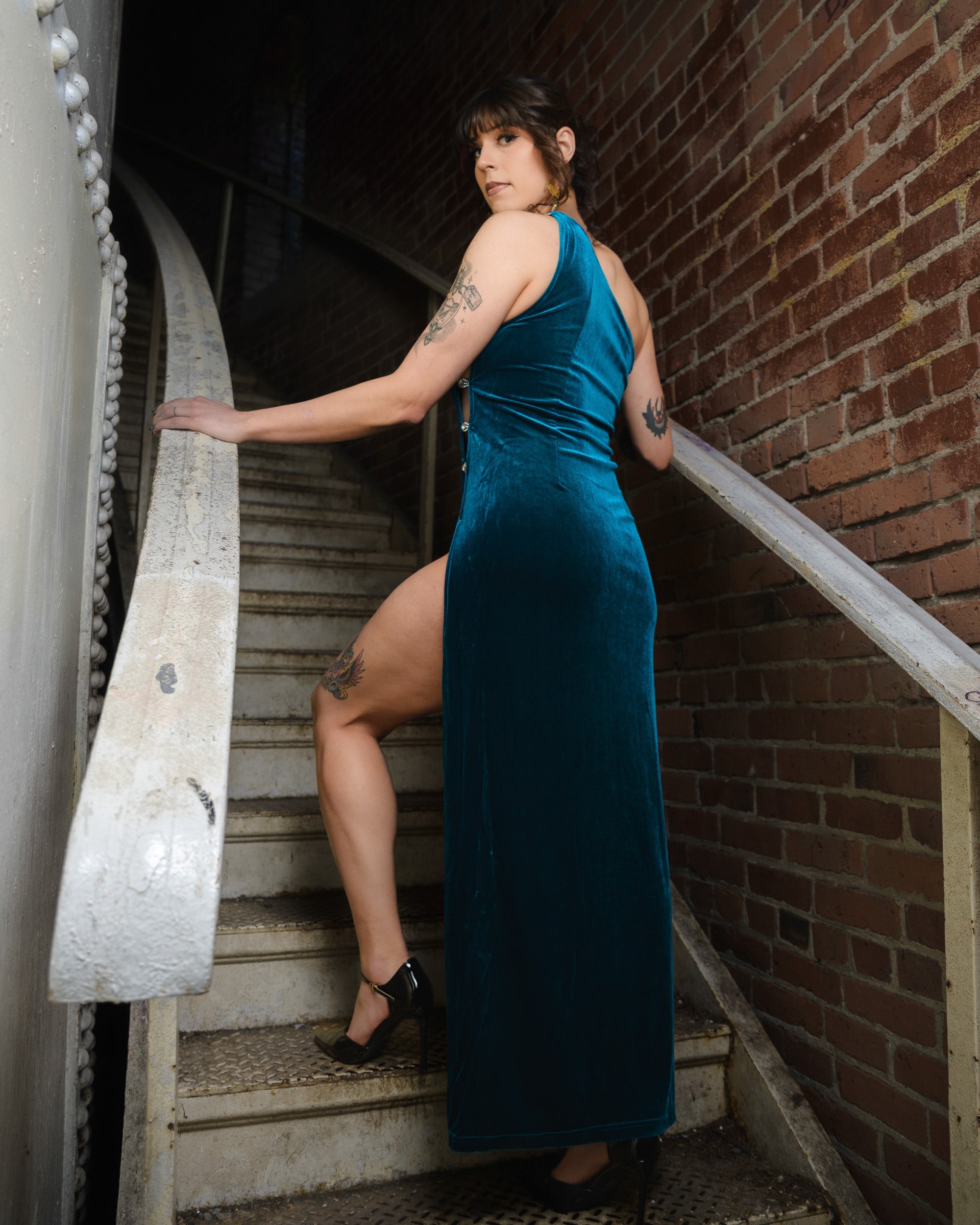 Woman in a teal dress standing on a staircase against a brick wall. [color_id:royalty-blue] [editorial]