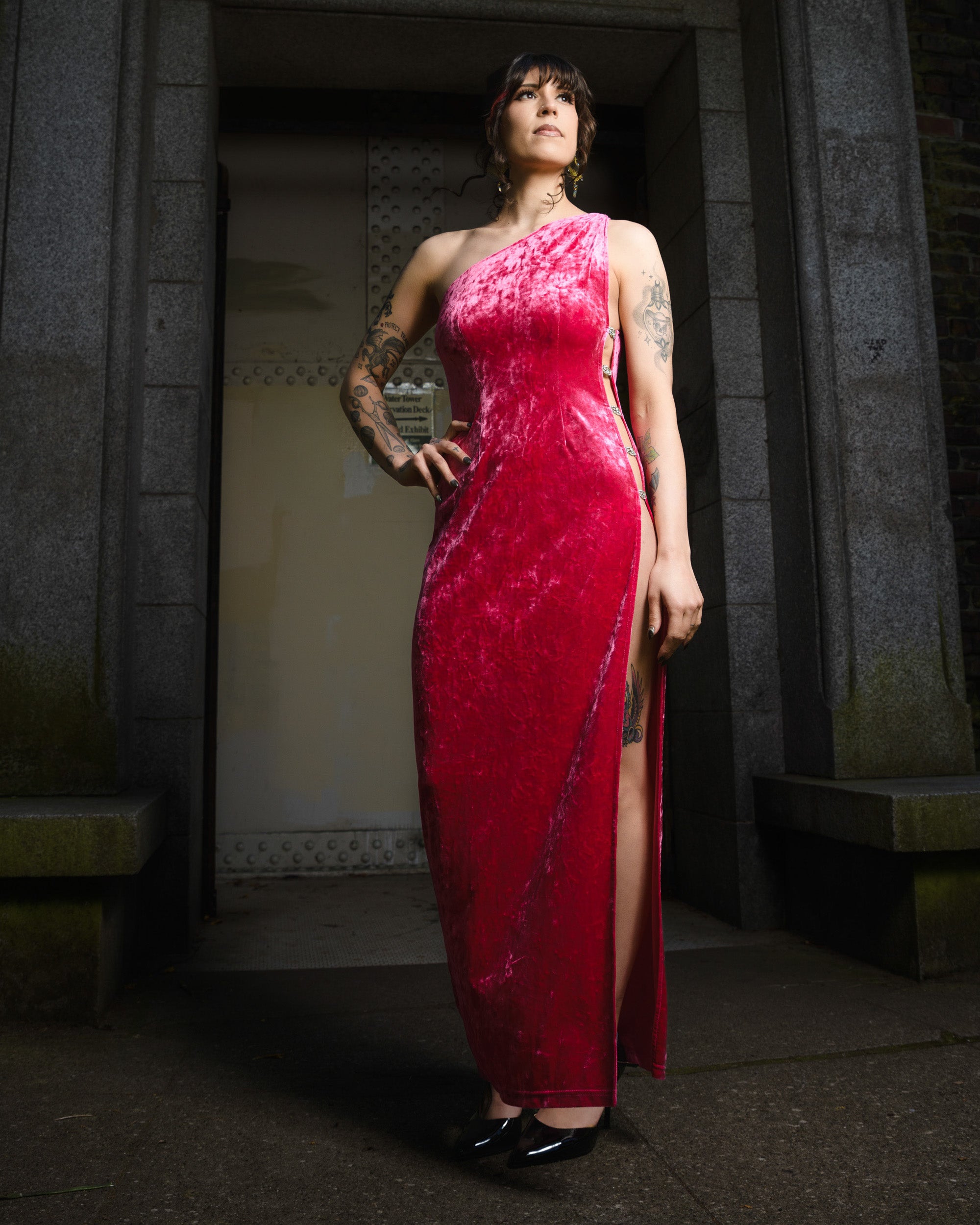 Woman in a red evening gown standing in a dimly lit hallway. [color_id:carnation-pink] [editorial] [model_size:M] 