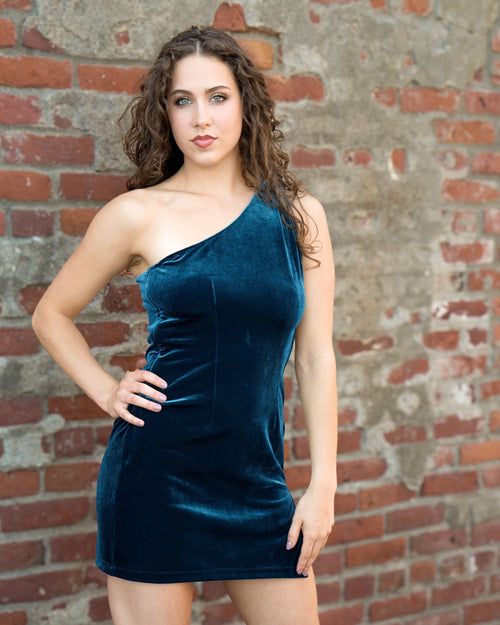 A woman modeling a short, one-shoulder velvet dress in a deep teal blue color, standing against a rustic red brick wall.   [style_size:M]