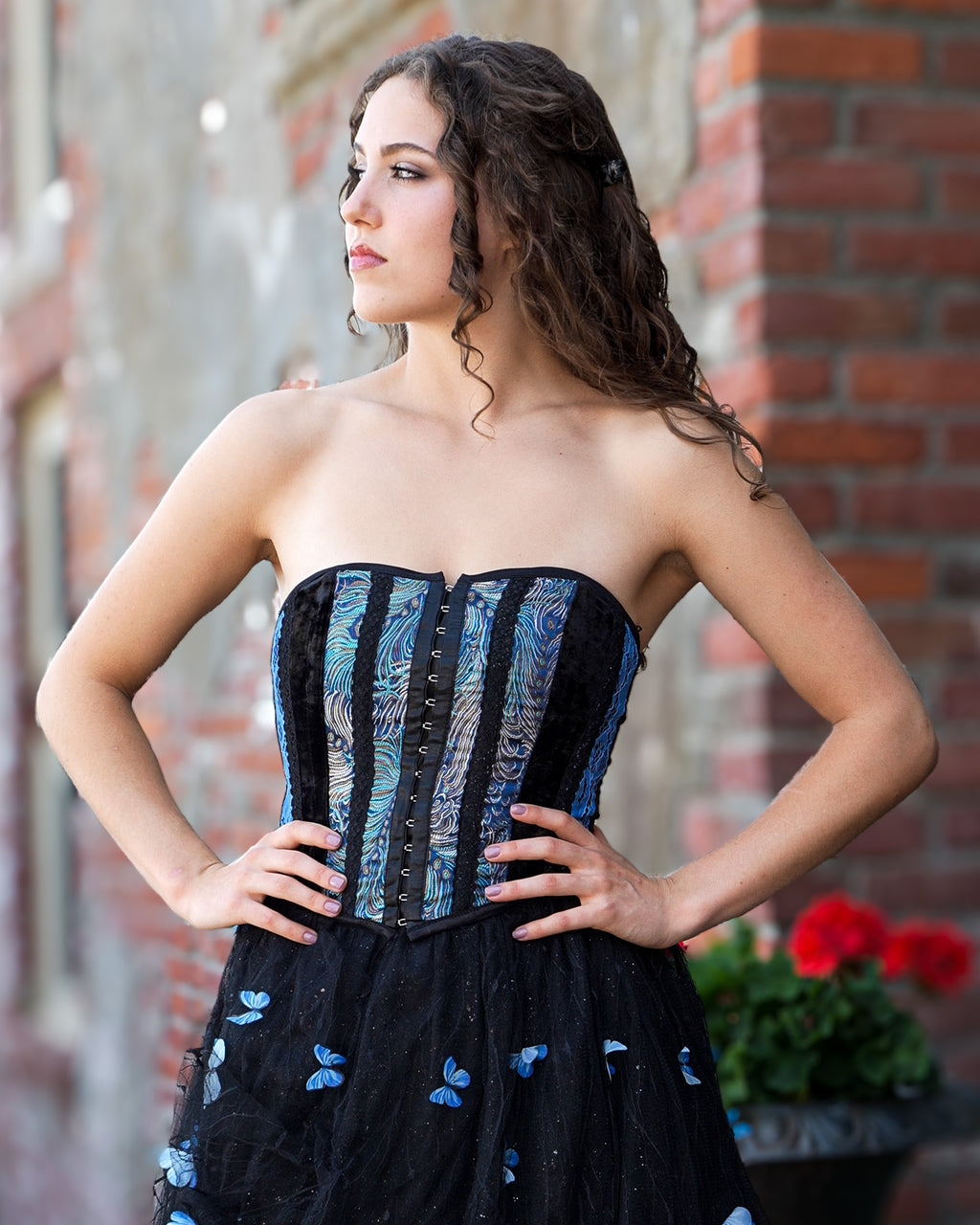A woman modeling a strapless black corset dress with blue patterned panels on the bodice and blue butterfly motifs on the skirt, posed outdoors against a brick wall.  [style_size:M]