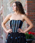 A woman modeling a strapless black corset dress with blue patterned panels on the bodice and blue butterfly motifs on the skirt, posed outdoors against a brick wall.  [style_size:M]