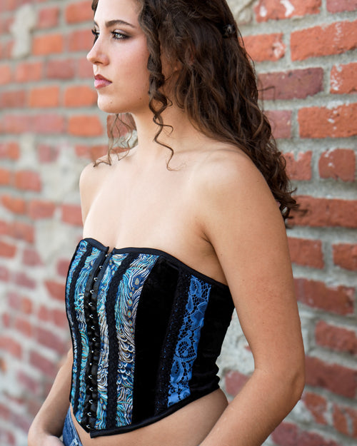 A woman wearing a strapless corset top with vertical panels of black velvet and blue lace-like patterns, standing against a red brick wall.   [style_size:M]