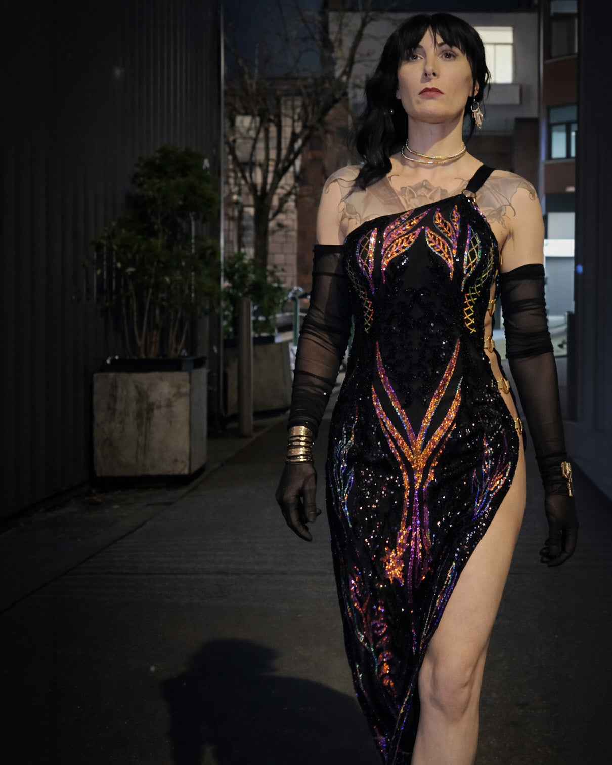 Woman in a black sequined dress with colorful patterns standing on a street at night. [color_id:black] [hero] 