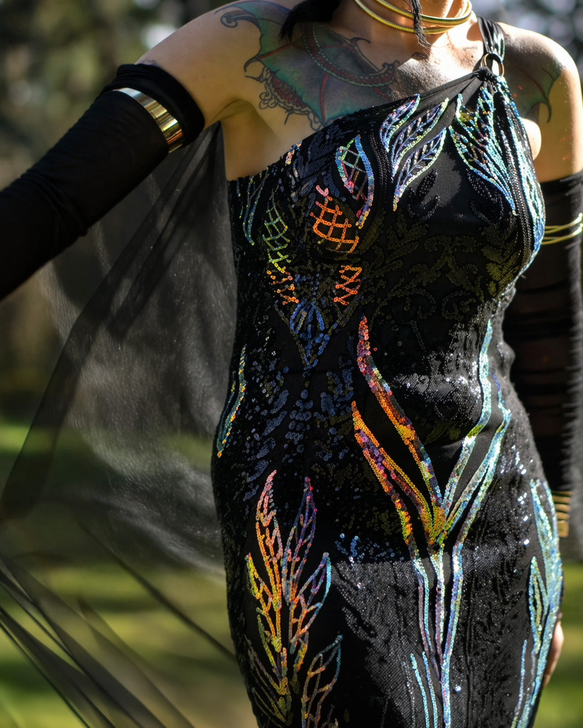 Person wearing a black sequin dress with colorful patterns, holding a sheer fabric. [color_id:black]